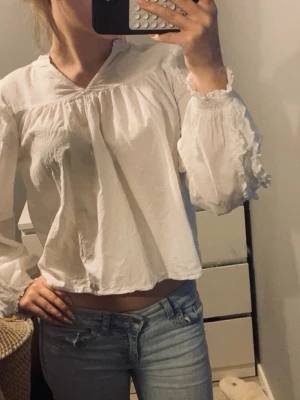 Vit bohemisk blus med volangärm - Luftig vit blus i bohemisk stil med v-ringning och volangdetaljer vid ärmslut. Blusen har en lös passform och är tillverkad i ett tunt, mjukt tyg som ger en avslappnad look. Perfekt att matcha med jeans för en trendig vardagsstil.