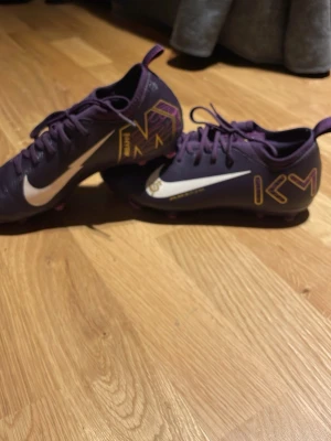 Lila Nike Mercurial fotbollsskor - Säljer ett par lila Nike Mercurial fotbollsskor med vita swoosh-loggor och gula KM-detaljer på sidorna. Skorna har fasta dobbar i rosa och är tillverkade i syntetmaterial för bra grepp och kontroll på planen. Perfekta för dig som vill sticka ut på fotbollsplanen.