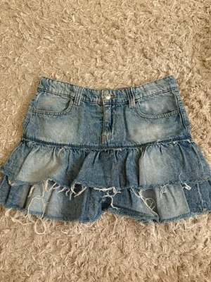 Blå denim skort med volanger - Cool blå denim skort med två lager volanger och slitna detaljer. Klassisk femficksdesign och knappstängning framtill. Materialet är jeans och färgen är ljusblå med slitningar för en trendig look.