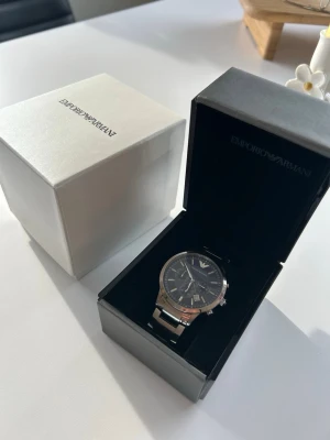 Emporio Armani silverklocka - Snygg klocka från Emporio Armani med silverfärgat armband i metall och mörkblå urtavla. Klockan har flera små urtavlor och klassisk design med logotypen på toppen. Perfekt för dig som vill ha en stilren accessoar.