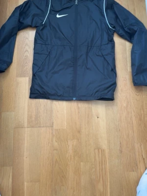 Svart Nike vindjacka med huva - Snygg svart vindjacka från Nike med vit logga på bröstet och vita detaljer längs axlarna. Jackan har dragkedja framtill, två sidofickor och huva med dragsko. Tillverkad i lätt och tåligt material som skyddar mot vind.