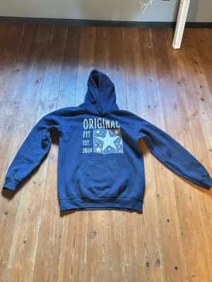 Blå hoodie med tryck och huva - Mörkblå hoodie från Original Fit med stor huva och tryck framtill med texten 'ORIGINAL FIT EST. 2024' och en stjärndesign. Klassisk känguruficka och ribbade muddar vid ärmslut och nederkant. Perfekt för en avslappnad streetstyle.