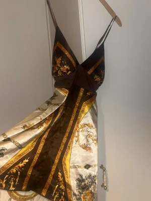Brun och guldig omlottklänning - Snygg kortklänning med omlottdesign i brunt, guld och beige. Klänningen har tunna axelband, v-ringning och ett elegant mönster med inslag av blommor och ornament. Materialet har en glansig satinlook som ger en lyxig känsla.