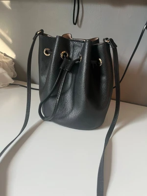 Svart bucket bag med gulddetaljer - Snygg svart bucket bag med justerbar axelrem och guldfärgade metalldetaljer. Väskan har dragskostängning och ljus insida, perfekt för dig som gillar stilrena accessoarer. Tillverkad i slätt konstläder som ger en modern look.
