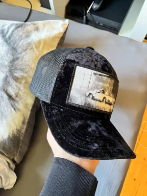Svart meshkeps med bilmotiv - Säljer en svart keps med mesh på sidorna och bak, samt en framsida i sammetsliknande material. På fronten finns en stor patch med ett svartvitt tryck av en sportbil. Kepsen har böjd skärm och justerbar snapback.