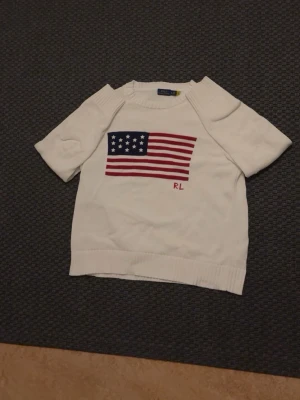Vit stickad tröja Ralph Lauren - Vit stickad tröja från Ralph Lauren med amerikansk flagga i blått, rött och vitt på bröstet samt RL-broderi. Tröjan har rund halsringning, ribbstickade muddar och lång ärm. Klassisk och stilren design som passar till mycket. Storlek Xl i barn storlek så passar runt M/L i vuxen storlek 