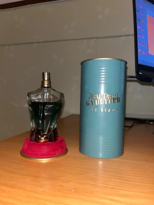 Jean Paul Gaultier Le Beau parfym - Unik parfymflaska formad som en manlig torso från Jean Paul Gaultier, Le Beau. Flaskan är gjord av genomskinligt glas med blågrön vätska och har en metallisk spraytopp. Medföljer gör en stilren turkos metalltub med gulddetaljer och logga.
