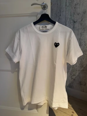Vit Comme des Garçons PLAY t-shirt - Vit t-shirt från Comme des Garçons PLAY med det ikoniska svarta hjärta-trycket på bröstet. Klassisk rund halsringning och korta ärmar. Tillverkad i mjuk bomull för en skön och avslappnad look. L men passar även M.