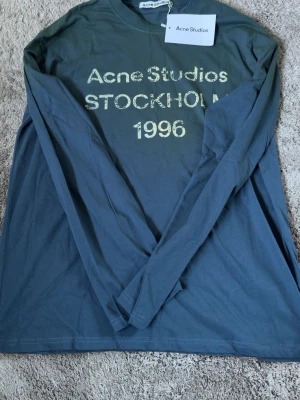 Acne Studios blå longsleeve  - Snygg blå långärmad topp från Acne Studios med tryck i vitt framtill: 'Acne Studios STOCKHOLM 1996'. Tillverkad i mjuk bomull och har en klassisk, normal passform. Perfekt för en clean och stilren look.