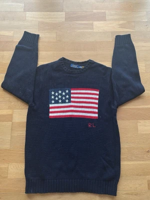 Mörkblå stickad tröja Polo Ralph Lauren - Mörkblå stickad tröja från Polo Ralph Lauren med amerikansk flagga i rött, vitt och blått på bröstet samt RL-broderi. Tröjan har rund halsringning, långa ärmar och ribbade muddar. Perfekt för dig som gillar klassisk preppy stil.