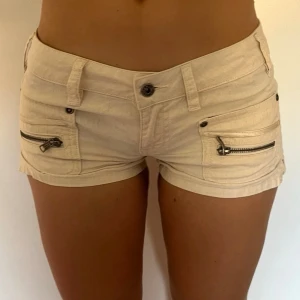 Beige/vita shorts från Diesel med dragkedjor - Säljer ett par snygga beige shorts från Diesel med låg midja. Shortsen har två framfickor med dragkedjor och bälteshällor. Materialet känns som bomull och de har en liten ficka bak med knapp och Diesel-logga. Perfekta för varma dagar!