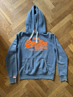 Grå Superdry hoodie med orange tryck - Snygg blå hoodie från Superdry med stort orange tryck på bröstet och japansk text. Klassisk känguruficka framtill, vita dragsnören i huvan och ribbade muddar vid ärmar och midja. Perfekt för en avslappnad streetwear-look.