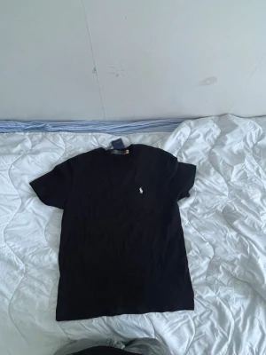 Svart t-shirt från Polo Ralph Lauren - Klassisk svart t-shirt från Polo Ralph Lauren med den ikoniska broderade loggan på bröstet. T-shirten har rund hals och korta ärmar, tillverkad i mjuk bomull för en skön känsla. Perfekt basplagg för en clean och stilren look.
