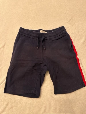 Mörkblå shorts med rödvit sidoremsa - Säljer ett par mörkblå shorts från pier one med snörning i midjan och en röd samt vit rand längs sidorna. Shortsen har en bakficka och är perfekta för sommaren. Sköna och stilrena för en avslappnad look.