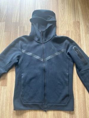 Svart Nike hoodie med dragkedja - Svart hoodie från Nike med hel dragkedja framtill och huva. En praktisk ficka med dragkedja på ena ärmen. Tillverkad i mjukt material som känns skönt mot huden. Perfekt för en sportig och avslappnad look.