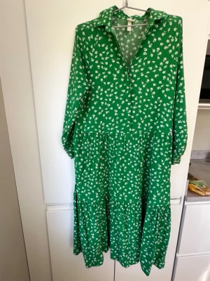 Grön blommig skjortklänning - Grön långärmad skjortklänning med vitblommigt mönster och krage. Klänningen har en avslappnad passform, knäppning framtill och volangdetaljer i kjolen. Perfekt för dig som gillar en färgglad och bekväm stil.