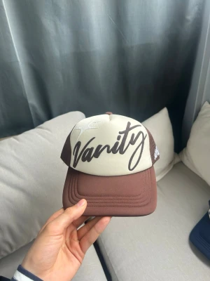 vanitywrld trucker keps - brun och krämvit truckerkeps från vanitywrld. Puff print logga på framsidan och embroidery text på sidan.