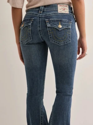 Blå bootcut jeans från True Religion - Snygga blå bootcut jeans från True Religion med klassiska kontrastsömmar och dekorativa fickor bak. Jeansen har låg midja, gylf med knapp och dragkedja samt detaljerade ficklock med logoknappar. Perfekta för en trendig och avslappnad look. 💕 lite slitage längst ner, som visas på sista bilden!!