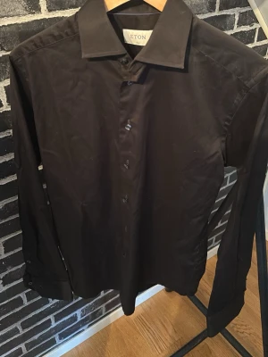 Svart skjorta från Eton  - Klassisk svart skjorta från Eton med lång ärm och knappar framtill. Skjortan har en stilren design med bred krage och är tillverkad i mjuk bomull som känns skön mot huden. Perfekt för dig som gillar en clean och tidlös look. Skriv om frågor 🤗