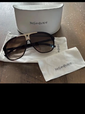 Yves Saint Laurent solglasögon - Snygga solglasögon från Yves Saint Laurent med svart ram och mörka glas. Modellen har en cool aviatorform med metallinslag upptill. Kommer med originalfodral och putsduk med logga. Perfekt för dig som vill ha en lyxig accessoar.