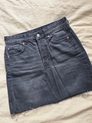 Svart jeanskjol med rå kant - Säljer en svart kort jeanskjol med rå, fransig kant nertill och klassiska fem fickor. Kjolen har knappgylf med synliga silverknappar och bälteshällor. Perfekt till en avslappnad och trendig look.
