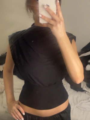 Svart croppad topp med markerade axlar - Säljer en svart croppad topp med markerade axlar och hög hals. Toppen har en tight passform i midjan och är ärmlös, vilket ger en cool och edgy look. Perfekt att styla med höga byxor eller kjol för en trendig outfit.
