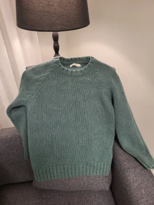Grön ull stickad tröja med rund hals TWOTHIRDS M - Mysig stickad tröja i dämpad grön färg med rund halsringning och ribbade muddar vid ärmslut och nederkant. Perfekt för kyliga dagar och enkel att matcha med jeans eller kjol.
