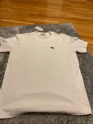 Vit t-shirt från Lacoste - Klassisk vit t-shirt från Lacoste med den ikoniska krokodilloggan broderad på bröstet. T-shirten har rund halsringning och korta ärmar. Tillverkad i mjuk bomull för en skön och enkel stil.