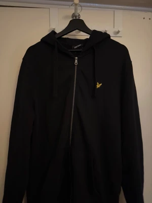 Svart hoodie med dragkedja Lyle & Scott - Säljer en svart hoodie från Lyle & Scott med dragkedja framtill och gul logga på bröstet. Hoodien har huva med dragsko och fickor framtill. Perfekt för en avslappnad och stilren look.