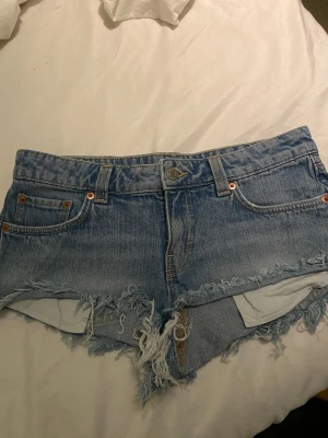Blå lågmidjade jeansshorts WEEKDAY - Snygga blå lågmidjade jeansshorts med fransig och sliten kant. Klassisk femficksmodell med knapp och dragkedja framtill. Perfekta för sommaren och ger en avslappnad vibe. Passar dig som gillar en lite råare stil.