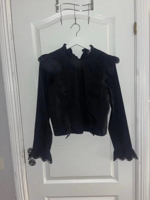 Svart blus med spetsdetaljer - Stilren svart blus med långa ärmar och dekorativa spetsdetaljer längs axlar, ärmslut och framsida. Helt ny med prislapp.