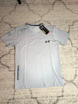 Ljusgrå träningströja från Under Armour - Snygg ljusgrå t-shirt från Under Armour med rund hals och korta ärmar. Tröjan har ett regnbågsskiftande UA-logotyp på bröstet och tryckt Under Armour-text längs sidan. Tillverkad i ett lätt och snabbtorkande funktionsmaterial, perfekt för träning.