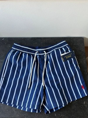 Randiga badbyxor från Polo Ralph Lauren - Snygga marinblå badbyxor från Polo Ralph Lauren med vita vertikala ränder och dragsko i midjan. Klassisk modell med fickor och röd logga broderad på benet. Perfekta för stranden eller poolen, tillverkade i snabbtorkande material.