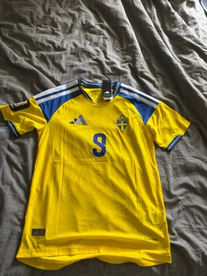 Sverige matchtröja Adidas nr 9 Isak - Gul och blå Sverige matchtröja med nummer 9 och namnet Isak på ryggen. Tröjan har korta ärmar, blå detaljer på axlar och ärmslut samt det svenska landslagets emblem på bröstet. Tillverkad i lätt och ventilerande material, perfekt för fotboll. Aldrig använd tröja därför 10/10 i skick. Vid frågor hör av er. Forza Sverige i Vm 