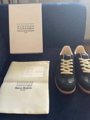 Maison Margiela svarta sneakers - Svarta sneakers från Maison Margiela med beige detaljer och vita skosnören. Ovandelen är i en mix av läder och mocka, med klassisk gummisula i ljusbrunt. Snygg och stilren design som passar till det mesta. Kommer med originalkartong och dustbag.