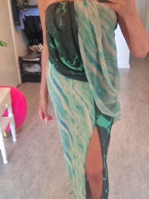 Grön mönstrad långklänning med slits - Säljer en snygg axelbandslös långklänning i gröna och ljusa toner med marmorerat mönster. Klänningen har en hög slits framtill och draperad detalj upptill. Perfekt för dig som vill sticka ut med en unik look.