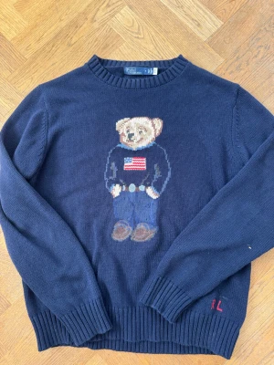 Mörkblå stickad tröja Polo Bear - Mörkblå stickad tröja från Polo Ralph Lauren med den ikoniska Polo Bear på bröstet, klädd i jeans och tröja med amerikanska flaggan. Tröjan har ribbade muddar och RL-broderi vid nederkanten. Perfekt för dig som gillar klassisk preppy stil. Nästan aldrig använd pga att den inte passar min stil!