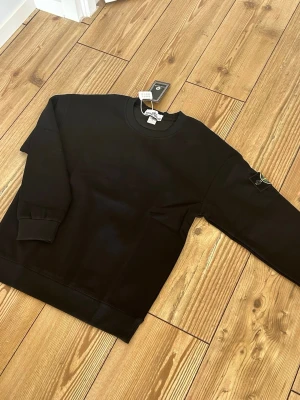 Svart sweatshirt från Stone Island - Snygg svart sweatshirt från Stone Island med klassisk rund halsringning och ribbade muddar. Tröjan har den ikoniska Stone Island-loggan på vänster ärm och är tillverkad i mjuk bomull. Perfekt för en clean och stilren look.