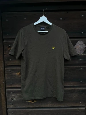 Olivgrön t-shirt från Lyle & Scott - Snygg olivgrön t-shirt från Lyle & Scott med klassisk gul logga på bröstet. T-shirten har rund halsringning och korta ärmar. Tillverkad i mjuk bomull för en bekväm känsla. Perfekt basplagg till garderoben.