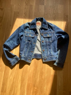 Klassisk blå jeansjacka - Snygg blå jeansjacka med raka linjer och klassisk krage. Två bröstfickor med lock och knappar framtill. Jackan har kontrastsömmar och metallknappar för en tidlös look. Perfekt att slänga över en hoodie eller t-shirt för en avslappnad stil.