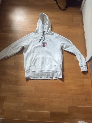 Vit hoodie från GANT med logga - Säljer en vit hoodie från GANT i storlek XL. Hoodien har en stor huva med dragsko, känguruficka framtill och GANTs klassiska logga med sköld på bröstet. Tillverkad i mjukt bomullsmaterial och har ribbade muddar vid ärmslut och nederkant.