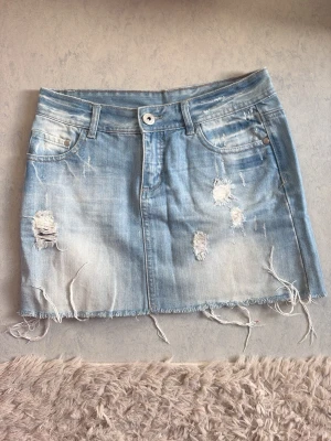 Ljusblå jeanskjol med slitningar - Säljer en ljusblå kort jeanskjol med slitna detaljer och fransig nederkant. Kjolen har klassiska fickor, väldigt fint skick, blivit för liten tyvärr, otroligt fin när man är lite brunare. Storleken är Xs/S