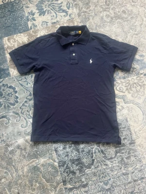Marinblå pikétröja från Polo Ralph Lauren storlek M - Säljer en klassisk marinblå pikétröja från Polo Ralph Lauren med vit broderad logga på bröstet. Tröjan har korta ärmar, krage och knappar framtill. Perfekt för dig som gillar stilrena och tidlösa plagg. Tröjan är helt ny och aldrig buren storlek M