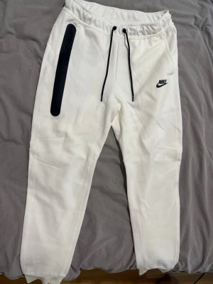 Vita mjukisbyxor från Nike - Säljer ett par vita mjukisbyxor från Nike med svart logga på vänster lår och svarta dragsnören i midjan. Byxorna har en svart detalj längs ena fickan och är tillverkade i ett mjukt, bekvämt material. Perfekta för en avslappnad stil.