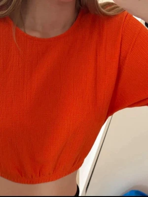 Croppad orange topp - Säljer en croppad topp i stark orange färg. Toppen har rund halsringning, korta ärmar och är tillverkad i ett strukturerat syntetmaterial. Perfekt för dig som vill sticka ut med en färgglad och trendig look.