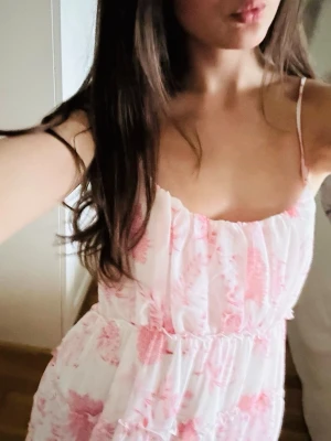 Vit och rosa blommig sommarklänning - Söt kortklänning i vitt med rosa blommigt mönster och tunna axelband. Klänningen har en luftig passform med volangdetaljer och är perfekt för varma dagar. Tillverkad i ett lätt och bekvämt material som känns skönt mot huden.
