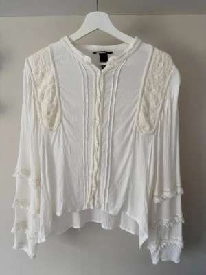 Vit boho-blus med spetsdetaljer - Superfin vit blus i boho-stil med spetsdetaljer på axlar och ärmar. Blusen har knappar framtill, lång ärm och lätt rynkat tyg som ger en luftig känsla. Perfekt till jeans eller kjol för en avslappnad look.