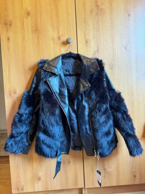 Svart skinnjacka med fuskpälsdetaljer - Snygg svart skinnjacka med breda fuskpälsdetaljer på ärmar och kropp. Jackan har klassisk krage, dragkedja och bälte i midjan för extra attityd. Perfekt för dig som vill sticka ut och addera lite edge till din outfit.