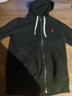 Svart hoodie från Polo Ralph Lauren - Säljer en svart hoodie från Polo Ralph Lauren med dragkedja, vit dragsko och klassisk röd logga på bröstet. Hoodien har känguruficka framtill och är tillverkad i mjukt bomullsmaterial. Perfekt för en avslappnad och stilren look.