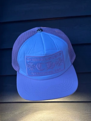 Ljusrosa Chrome Hearts keps - Säljer en ljusrosa truckerkeps från Chrome Hearts med broderad logga och text framtill. Kepsen har mesh på sidorna och bak, samt en klassisk böjd skärm. Märkets ikoniska detaljer och metallknapp på toppen ger en unik look.